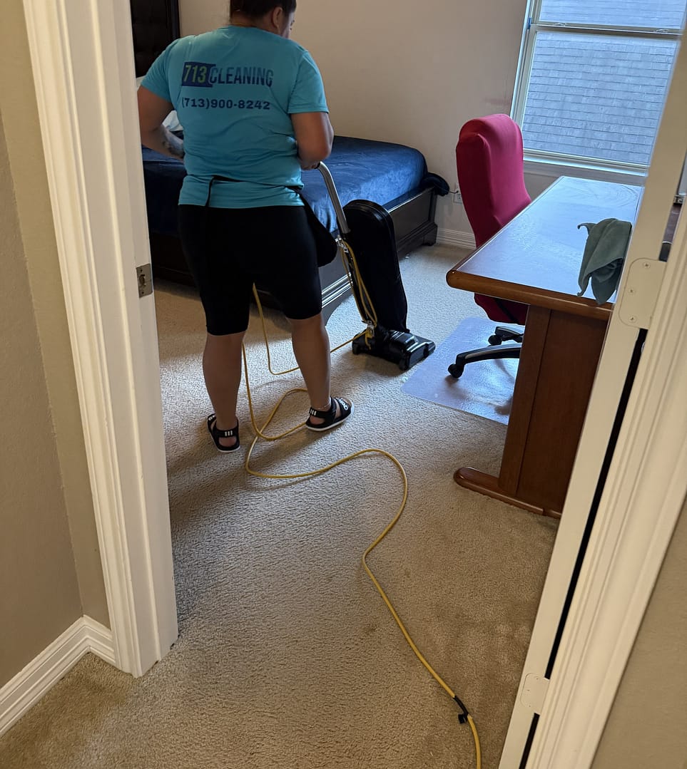 713 Cleaning team member vacuuming the carpet in a Houston-area bedroom, upright vacuum in front of her and the cord running back toward the doorway.