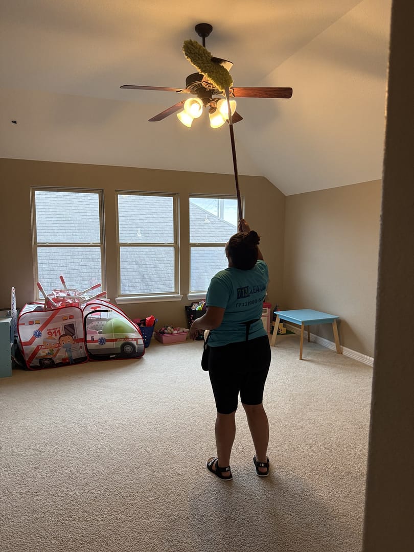 713 Cleaning team member using an extension duster to clean the blades and light fitting of a ceiling fan in a Houston-area playroom, toys and a low table behind her.