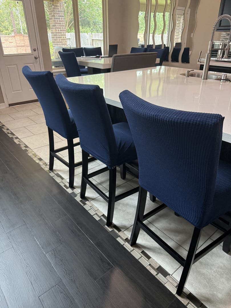 Kitchen island with a white quartz top and three navy upholstered bar stools, a dining table set behind it and a tiled floor meeting dark wood planks.