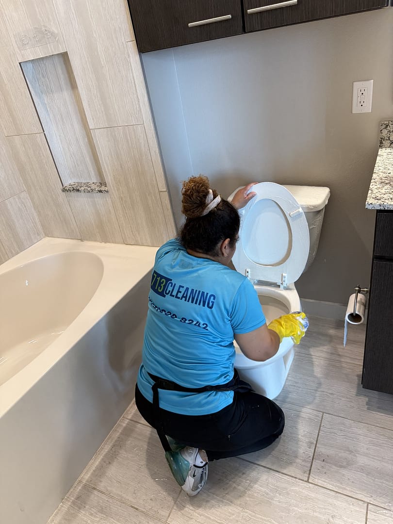 A 713 Cleaning crew member, seen from behind, kneels on a tile floor in yellow gloves and wipes the lifted seat and lid of a toilet beside a bathtub.