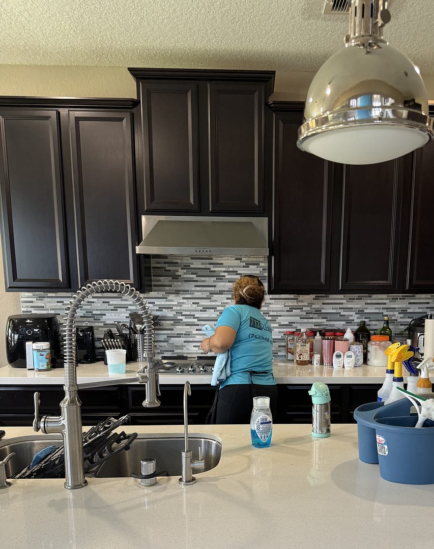 A 713 Cleaning crew member works at a back counter under a stainless range hood and dark wood cabinets, with stove grates set in the island sink in the foreground.