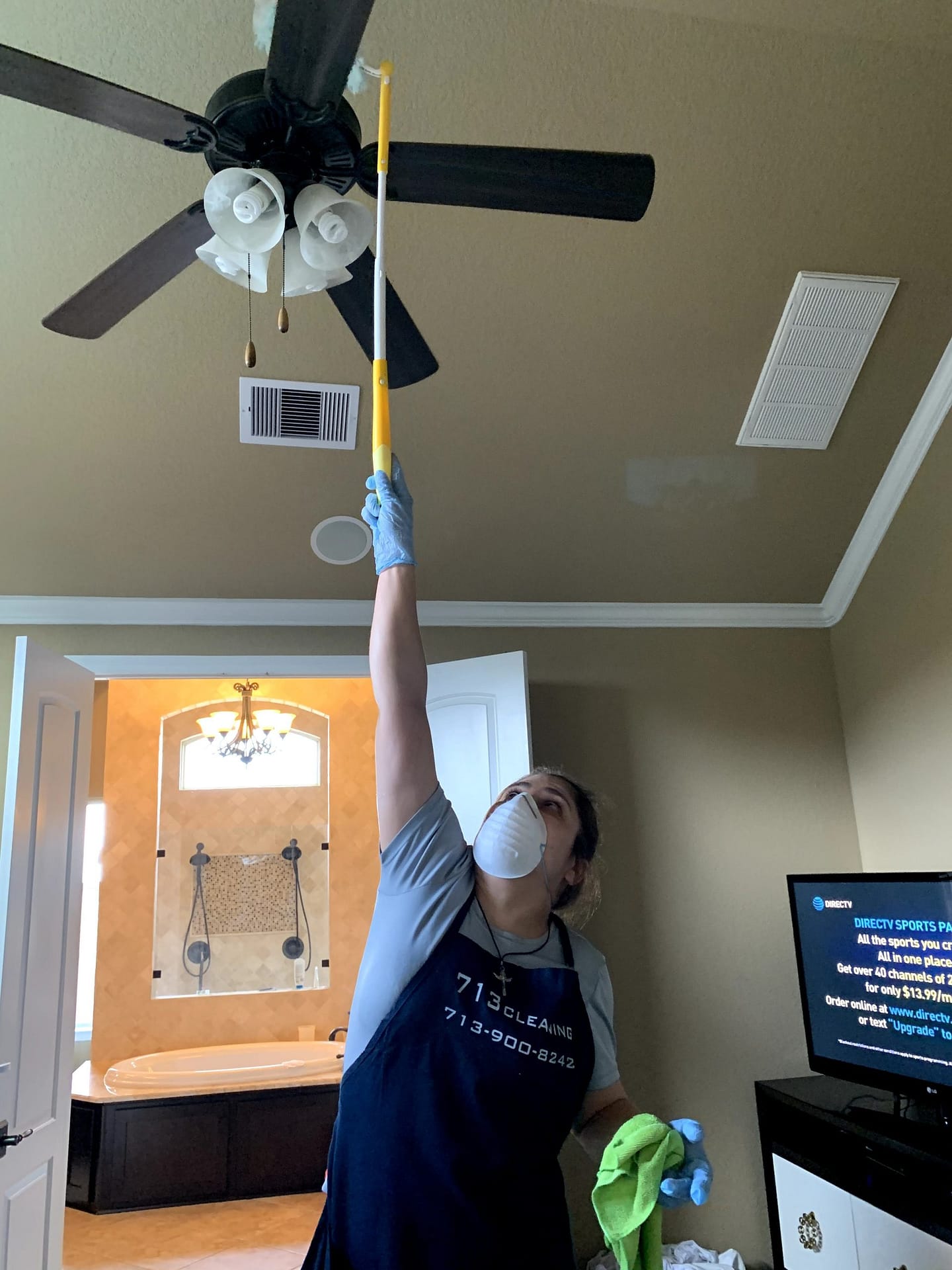 Uniformed 713 Cleaning crew member in gloves and mask dusting a ceiling fan in a client home.