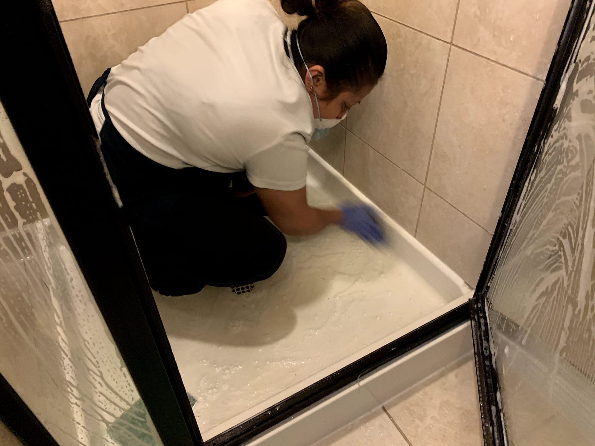 Cleaning crew member in gloves and a mask hand-scrubbing a lathered, tiled shower floor.