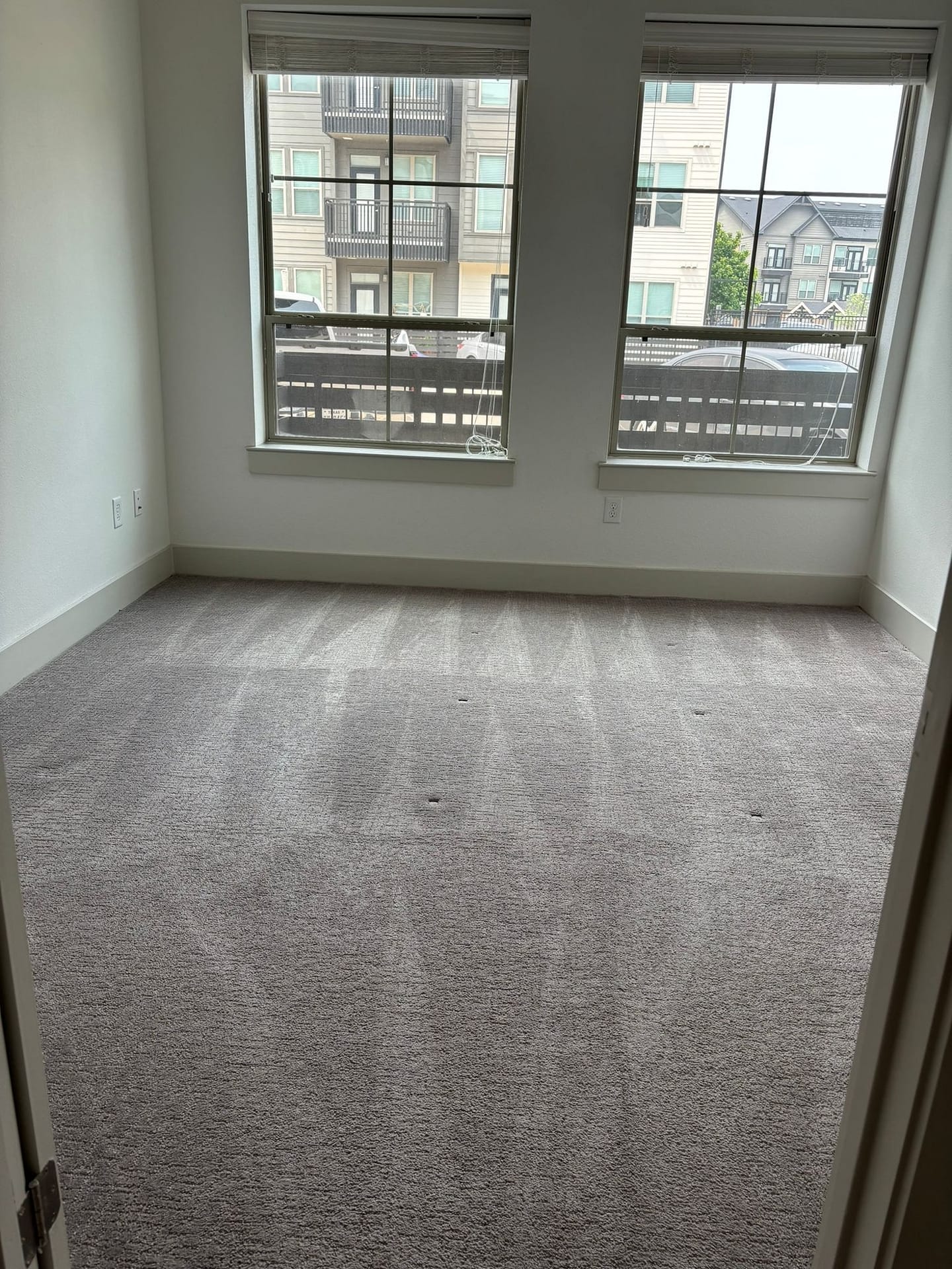 Empty carpeted bedroom with fresh overlapping vacuum passes visible in the pile, white baseboards and two windows.
