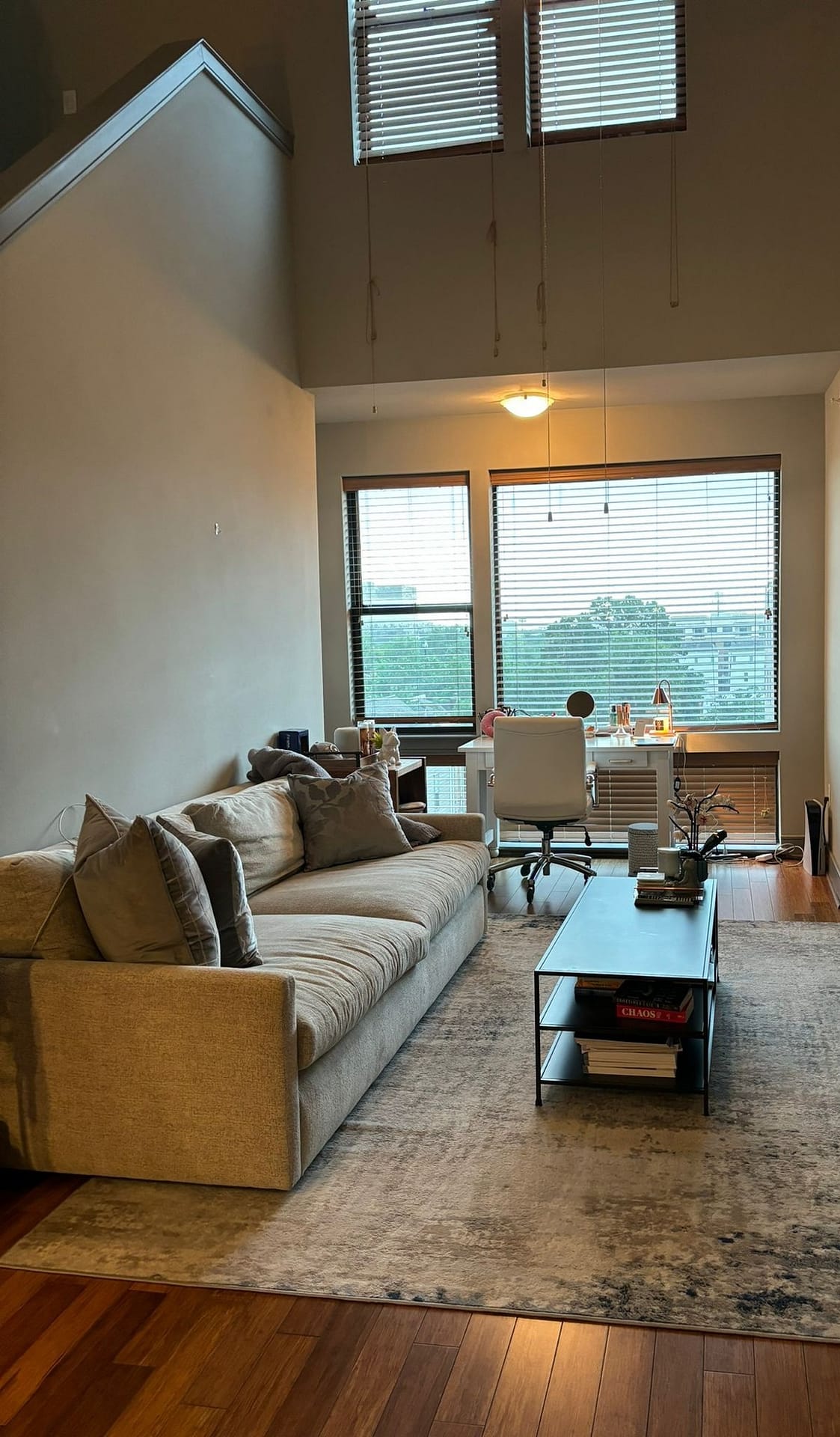 Living room with a beige fabric sofa and gray cushions on a patterned rug over a wood floor, a desk and tall windows behind it.