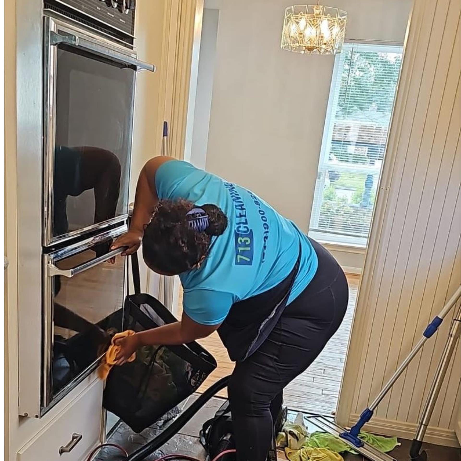 713 Cleaning crew member wiping down the open door and interior edge of a double wall oven in an empty Houston home, with the vacant dining room behind.