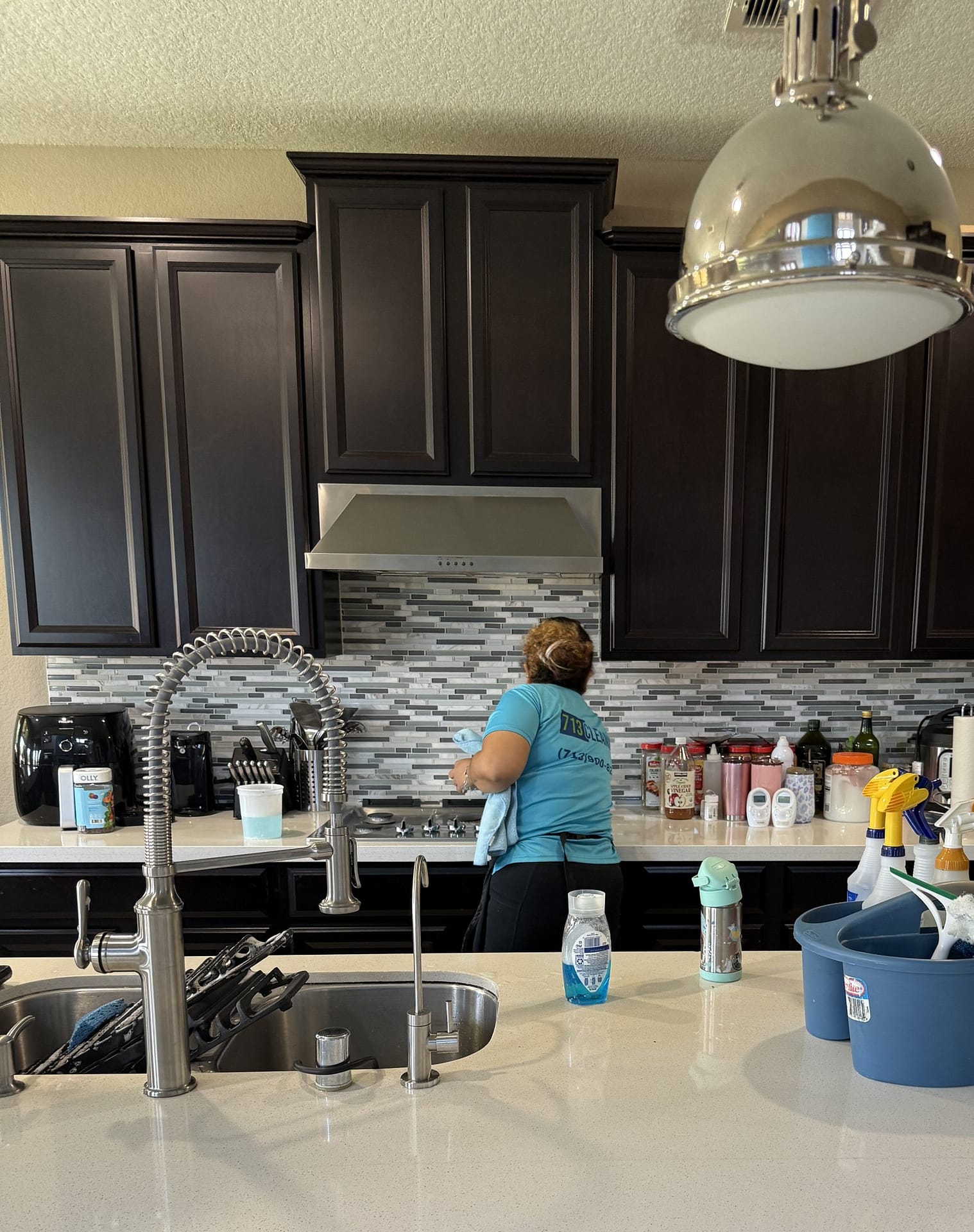 A 713 Cleaning crew member works at a back counter under a stainless range hood and dark wood cabinets, with stove grates set in the island sink in the foreground.